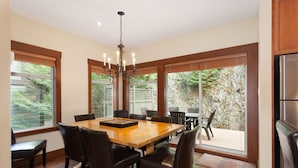 Un coin repas avec une table et des chaises en bois, de grandes fenêtres équipées de stores et un lustre.