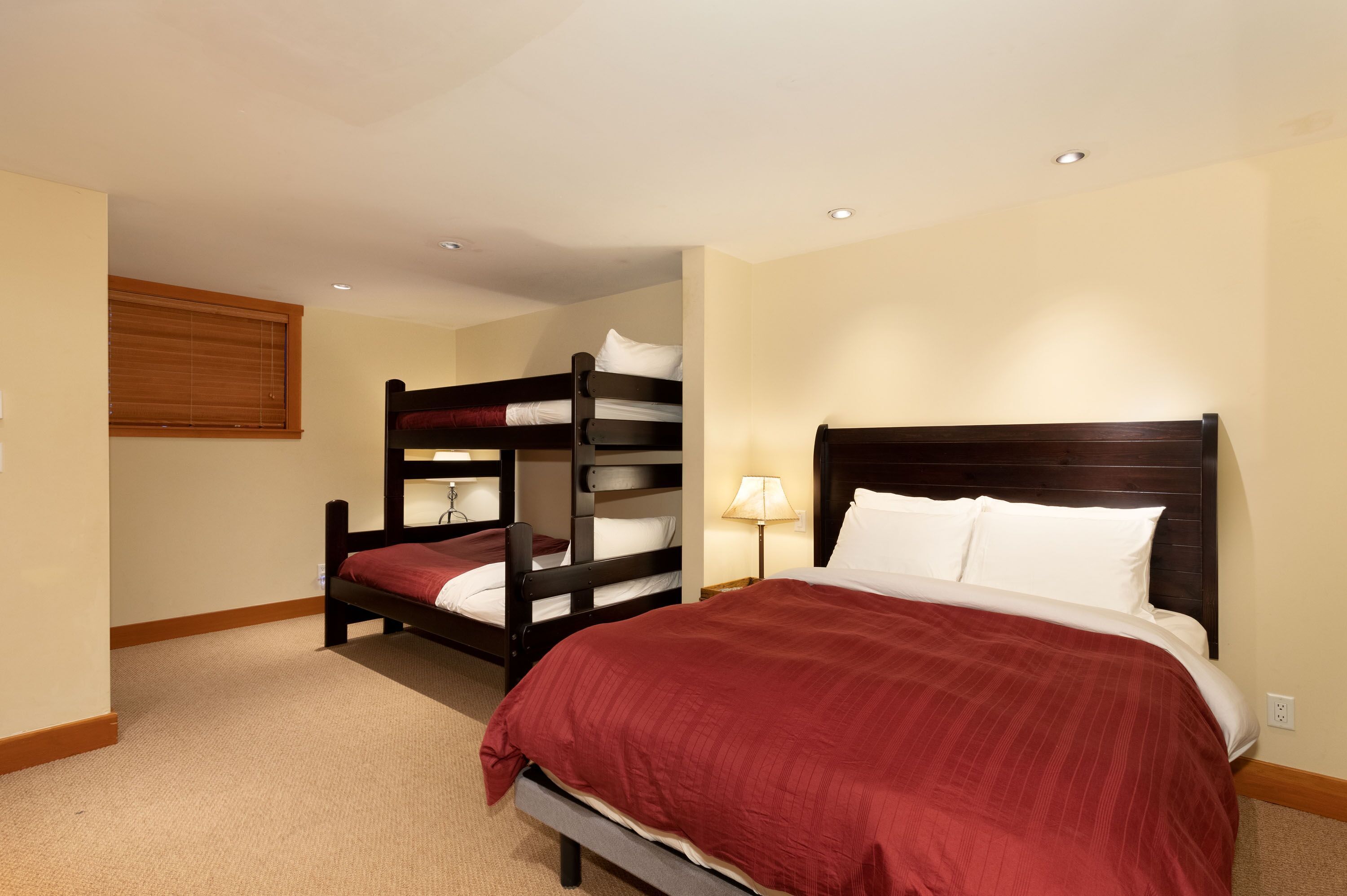 A room with a large bed, a smaller bed, and a bunk bed. There is a window with blinds, a lamp, and a ceiling light.