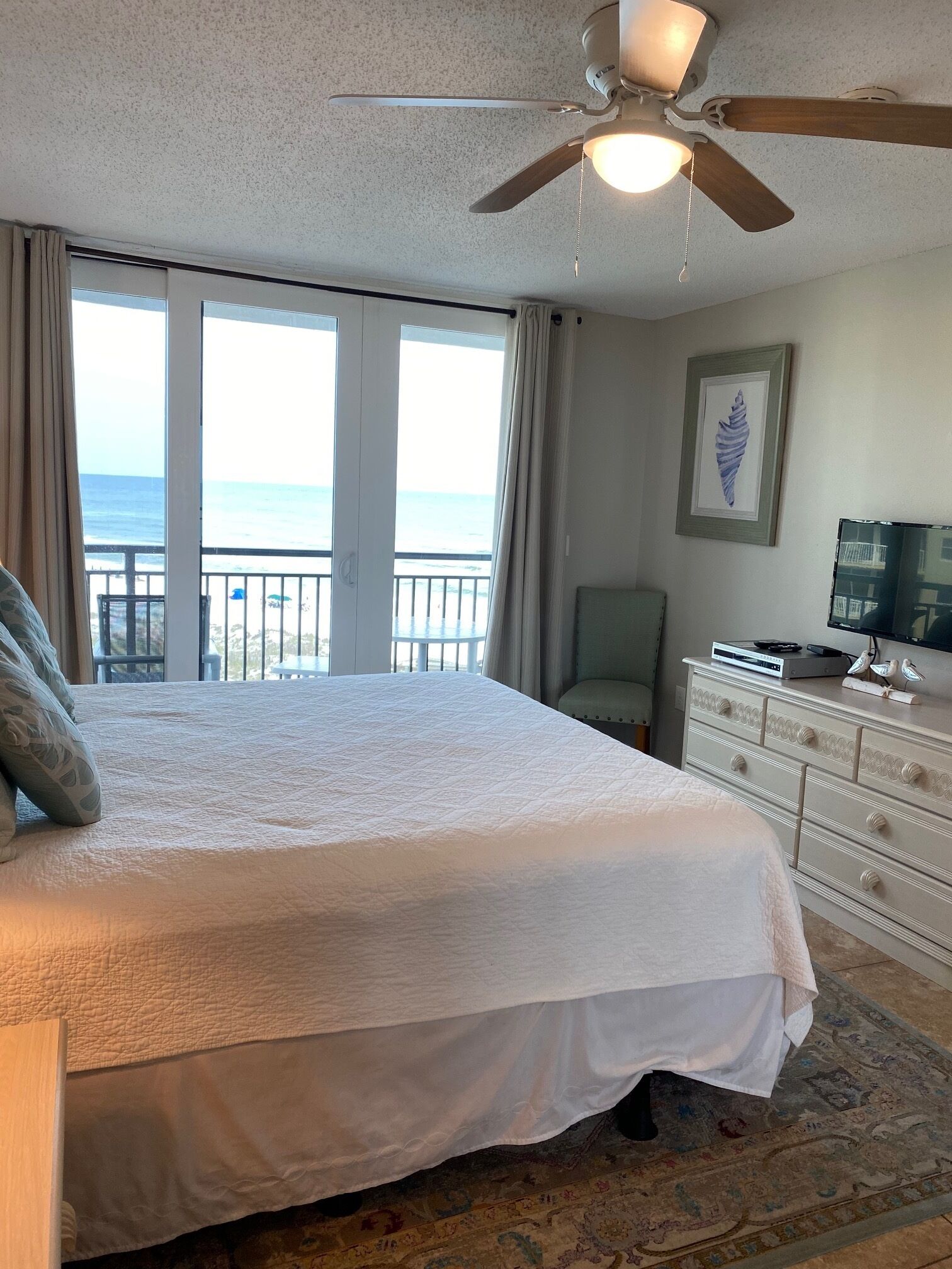 A bedroom with a large bed, a ceiling fan, a dresser, a chair, and a view of the ocean through large windows.