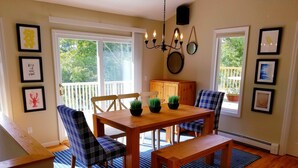 Un coin repas avec une table en bois, deux chaises, un banc et un lustre. Les murs sont ornés de tableaux encadrés, et les fenêtres offrent une vue sur l’extérieur.