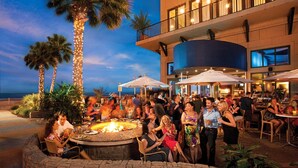 An outdoor dining area with a fire pit, surrounded by palm trees and umbrellas, overlooking a beach.