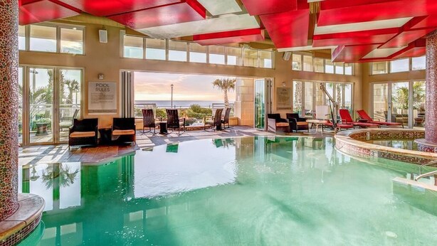A large indoor swimming pool with clear glass walls, red ceiling panels, and multiple lounge areas with chairs and sun loungers.