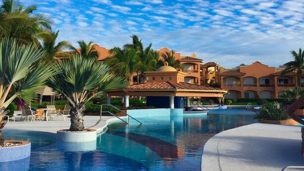 A resort pool area with a clear blue sky, palm trees, and a building with a tiled roof. - 2 BR condo nestled between Pacific Ocean beach and golf course    (Mazatlan)