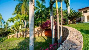 A landscaped garden with palm trees, a stone wall, and a building in the background. - Luxury Villa, Panoramic Views, Infinity Pool & Concierge (Punta Mita)