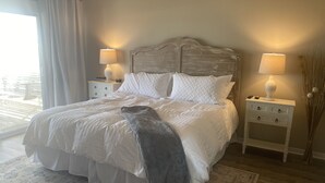 A neatly made bed with white bedding and a gray throw blanket, flanked by two bedside tables with lamps, in a room with a large window and a patterned rug.