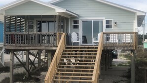 A beachfront house with a wooden deck, a staircase, and a large sliding glass door.