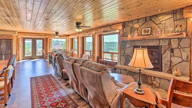 A spacious room with a stone fireplace, leather seating, and large windows offering a view of the outdoors. - House - Southern Hills Serene Getaway (Hot Springs)