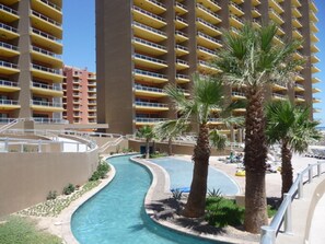 A swimming pool with a curved design, surrounded by palm trees and a high-rise building with yellow balconies in the background. - Las Palomas 3 BR/3BA 360/485 Stay 7 nights get one free (Puerto Penasco)