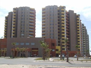 A high-rise residential building with multiple balconies, a central entrance, and a landscaped area with palm trees. - Las Palomas 3 BR/3BA 360/485 Stay 7 nights get one free (Puerto Penasco)