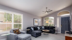 A modern living room with a grey sectional sofa, a glass coffee table, and a small side table. There is a ceiling fan, a framed artwork on the wall, and a door with a window view. - Sun, Fun, Golf, Swim, Relax in Your Own Resort Home. (Glendale)