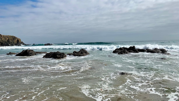 Un rivage rocheux où les vagues s’écrasent contre les rochers, sous un ciel partiellement nuageux.