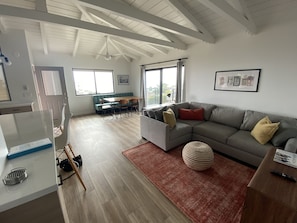Relax in a spacious living area with a high ceiling and exposed beams, featuring a comfortable sofa, a round ottoman, and a dining area with a bench and table.