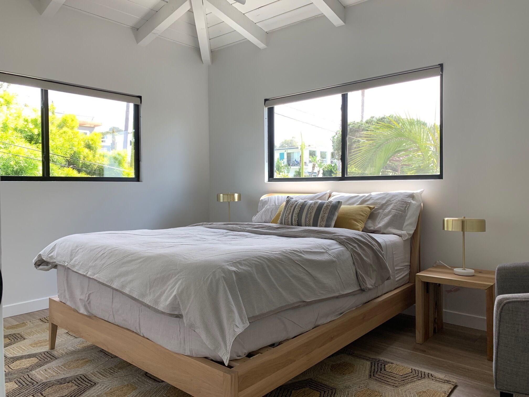 A modern bedroom with a wooden bed, two bedside lamps, and large windows offering a view of greenery and buildings.