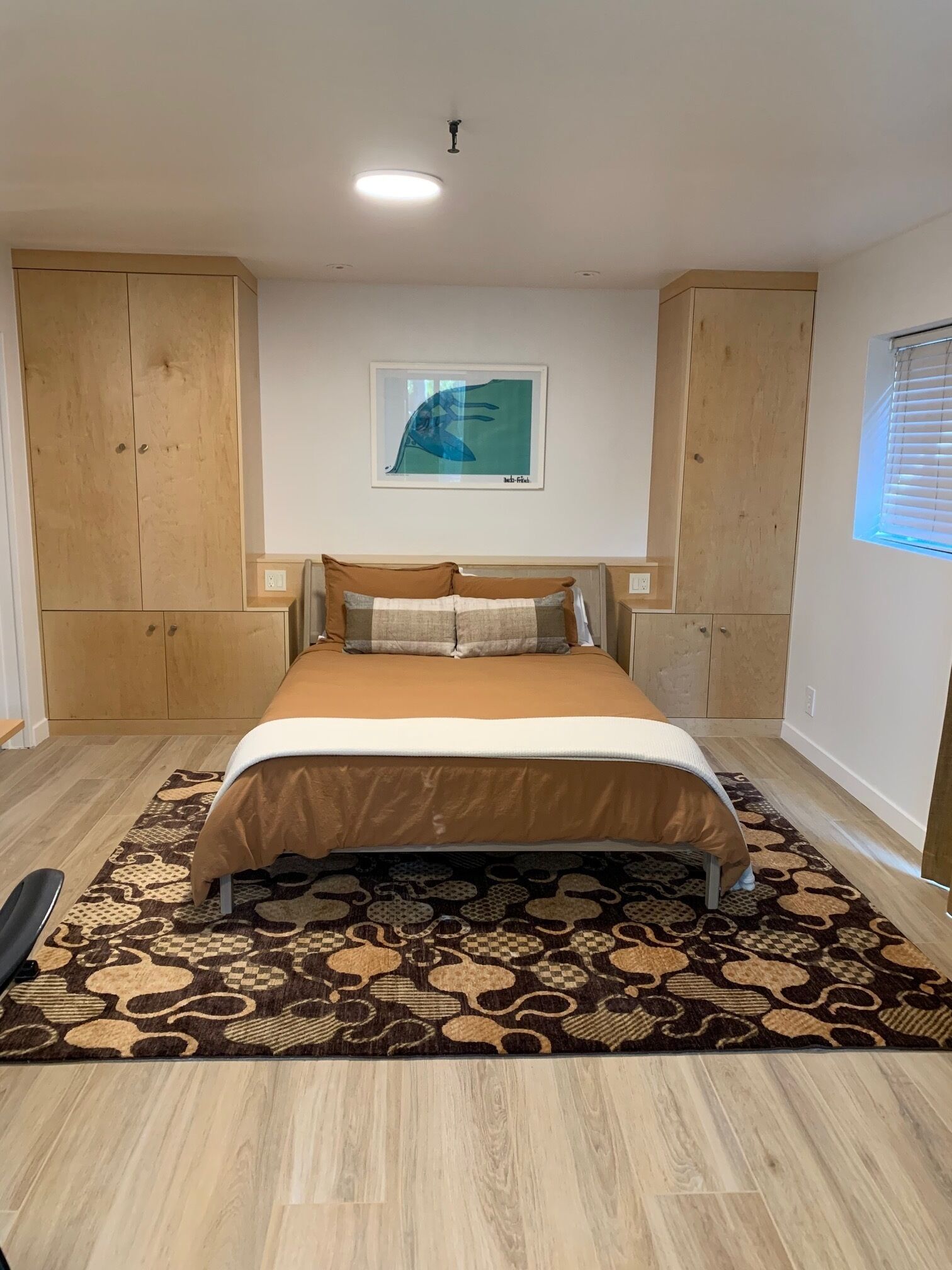 A bedroom with a bed, wooden wardrobe, a framed picture on the wall, and a window with blinds.
