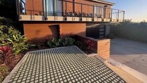 Una casa moderna de dos pisos con balcón, entrada pavimentada y jardín con plantas.