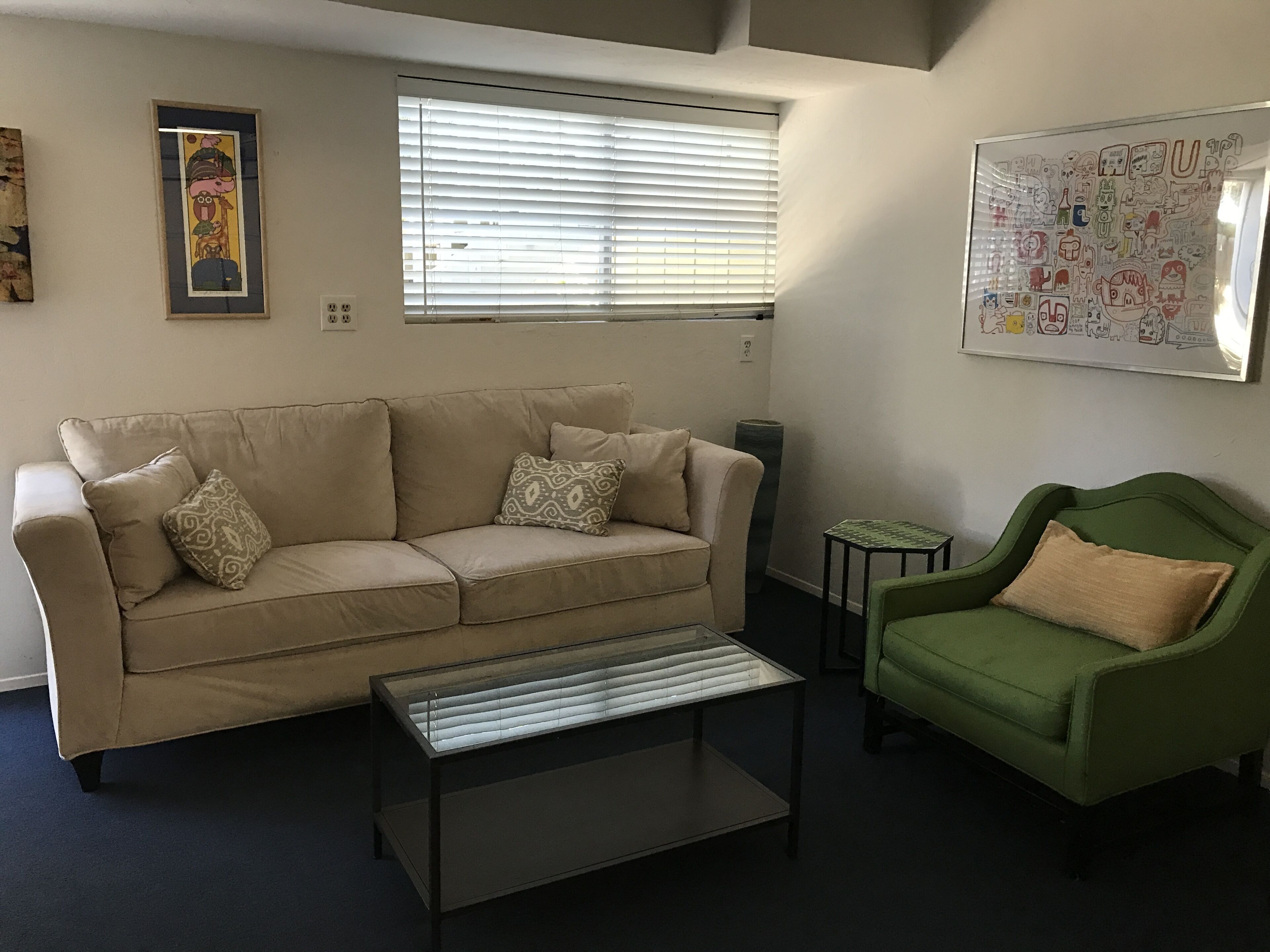 A living room with a beige sofa, a green armchair, a glass coffee table, and a window with blinds.