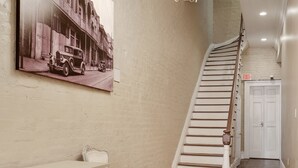 A hallway with a wooden staircase, a chandelier, a patterned rug, a white bench, and a vintage car painting on the wall. - Luxury 2-Story Condo / Prime Location (New Orleans)