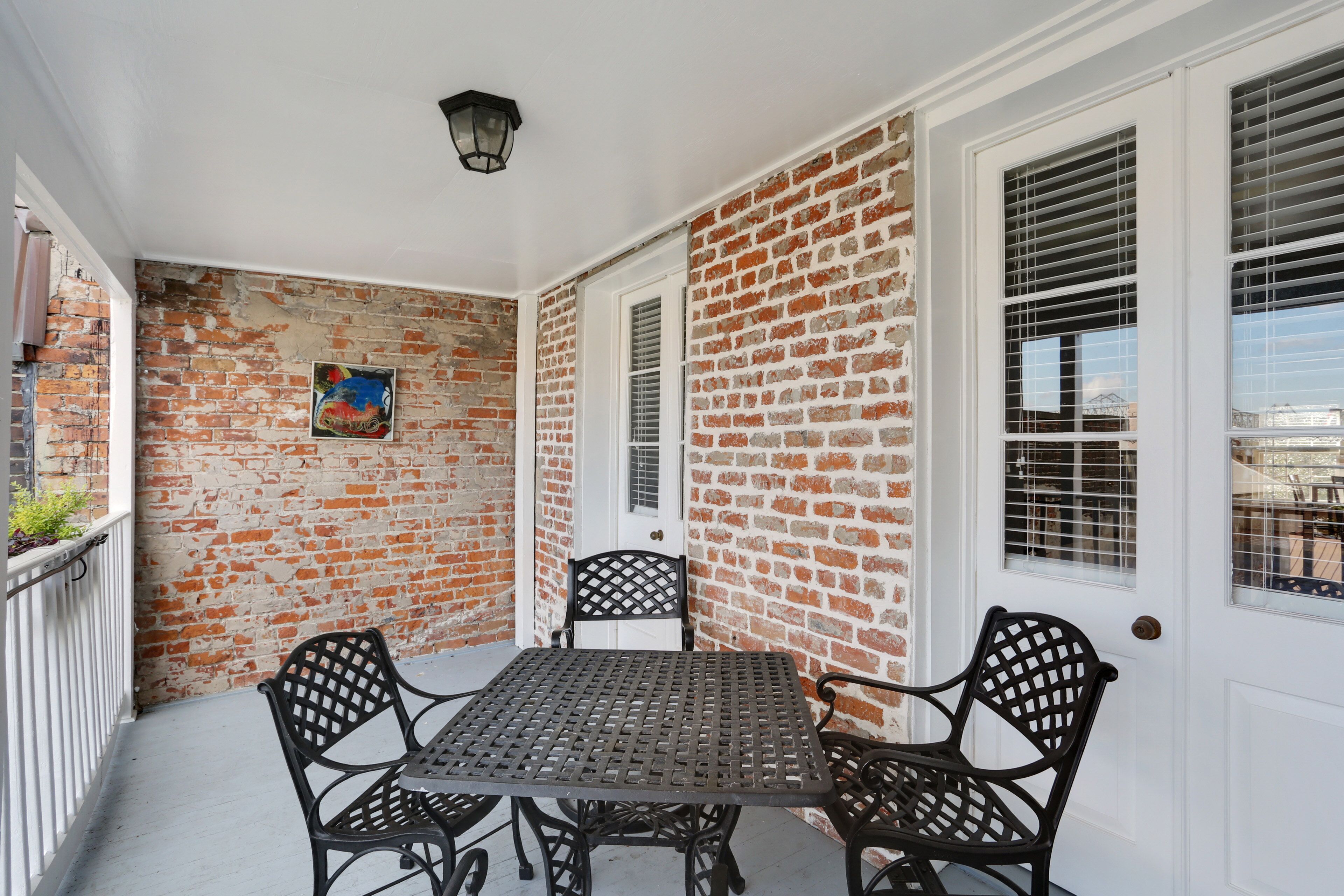 A brick wall with a white door and windows, a metal table with four chairs, and a small painting on the wall.