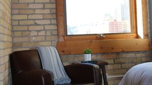 A room with a brick wall, a leather chair, a small table with a plant, a window with a view of buildings, and a bed with white bedding.