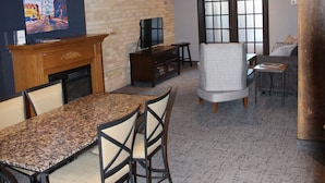 A dining area with a granite table and chairs, a fireplace, a television, and a large window with blinds.