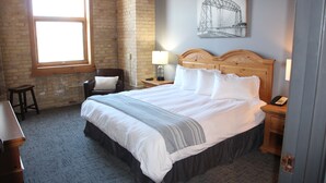 A hotel room with a large window, a bed with white linens, a wooden headboard, a nightstand with a lamp, and a chair.