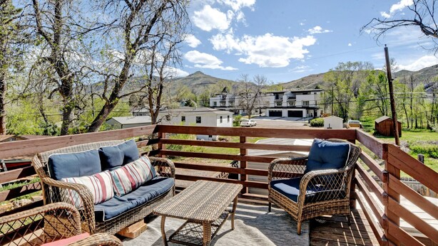 Une terrasse en bois, meublée de meubles en osier, avec une table et une vue sur une montagne et des bâtiments modernes en arrière-plan.