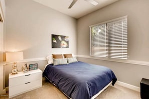Quarto com cama, mesa de cabeceira, janela com persianas e uma obra de arte na parede.