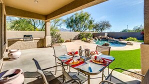 Un patio exterior con una mesa lista para comer, una parrilla de barbacoa y una piscina al fondo.