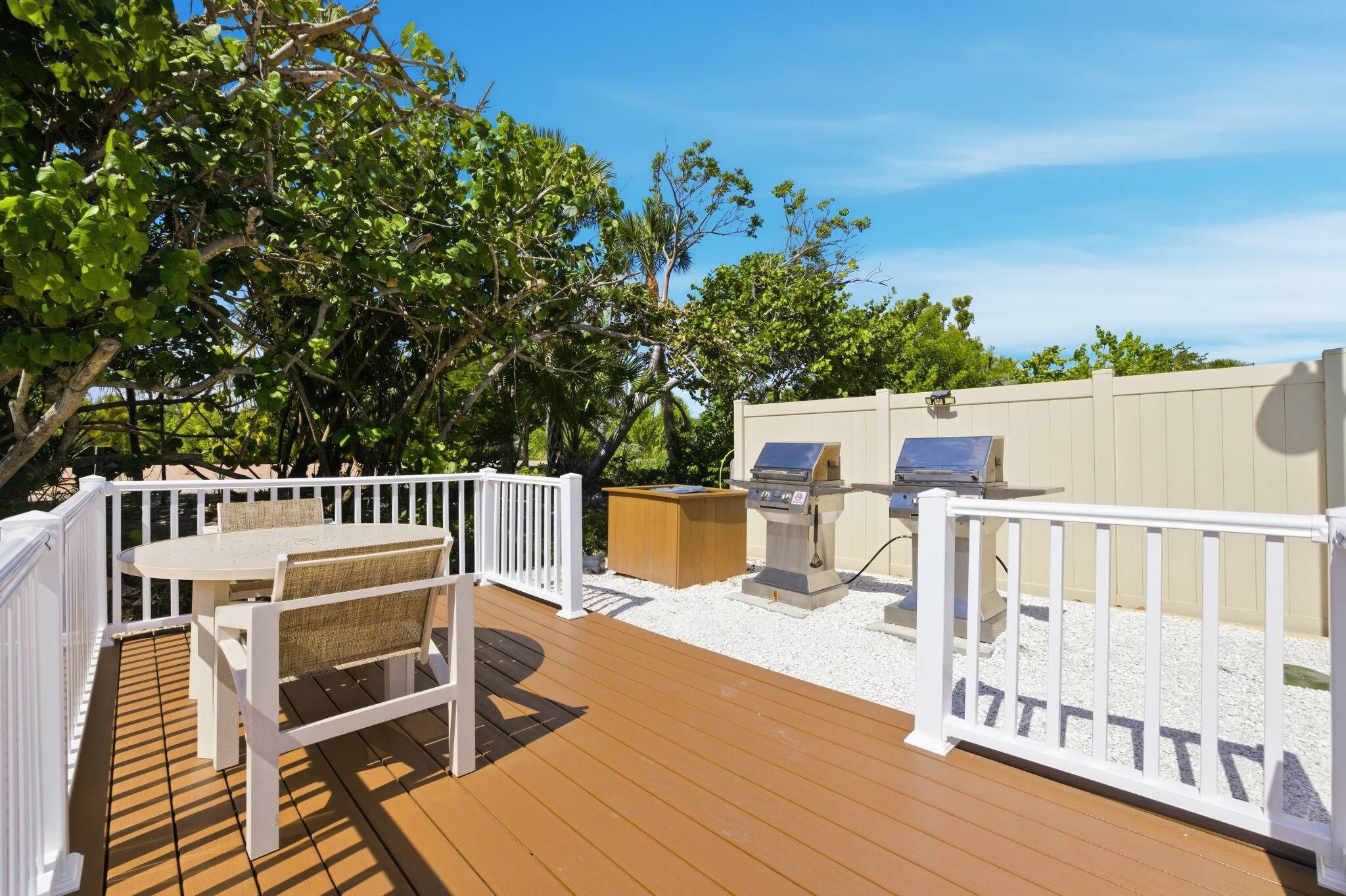 A wooden deck with a white railing, a table with chairs, and two barbecue grills.