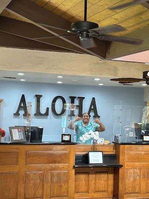 Uma mesa de recepção com um grande letreiro 'ALOHA', um ventilador de teto e painéis de madeira.