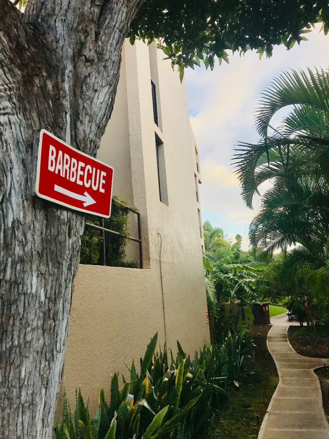 Un panneau indiquant un espace barbecue, un bâtiment à la façade beige et un sentier bordé de verdure.