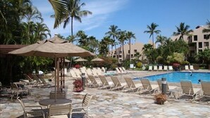 A poolside area with lounge chairs and umbrellas, surrounded by palm trees and a building in the background.