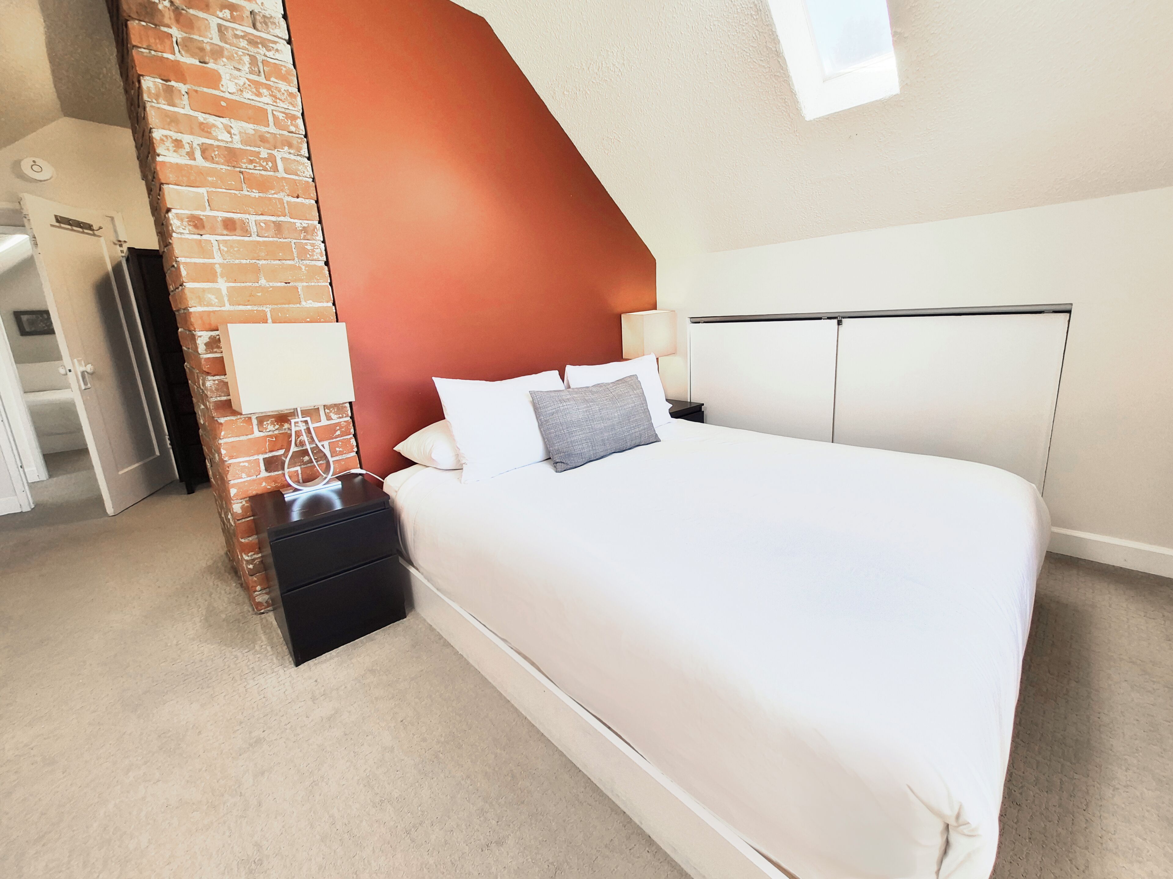 A bedroom with a brick accent wall, a white bed, a nightstand, a lamp, and a wardrobe.