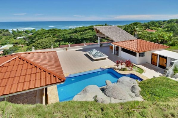 A modern house with a swimming pool, a thatched-roof structure, and a view of the ocean.