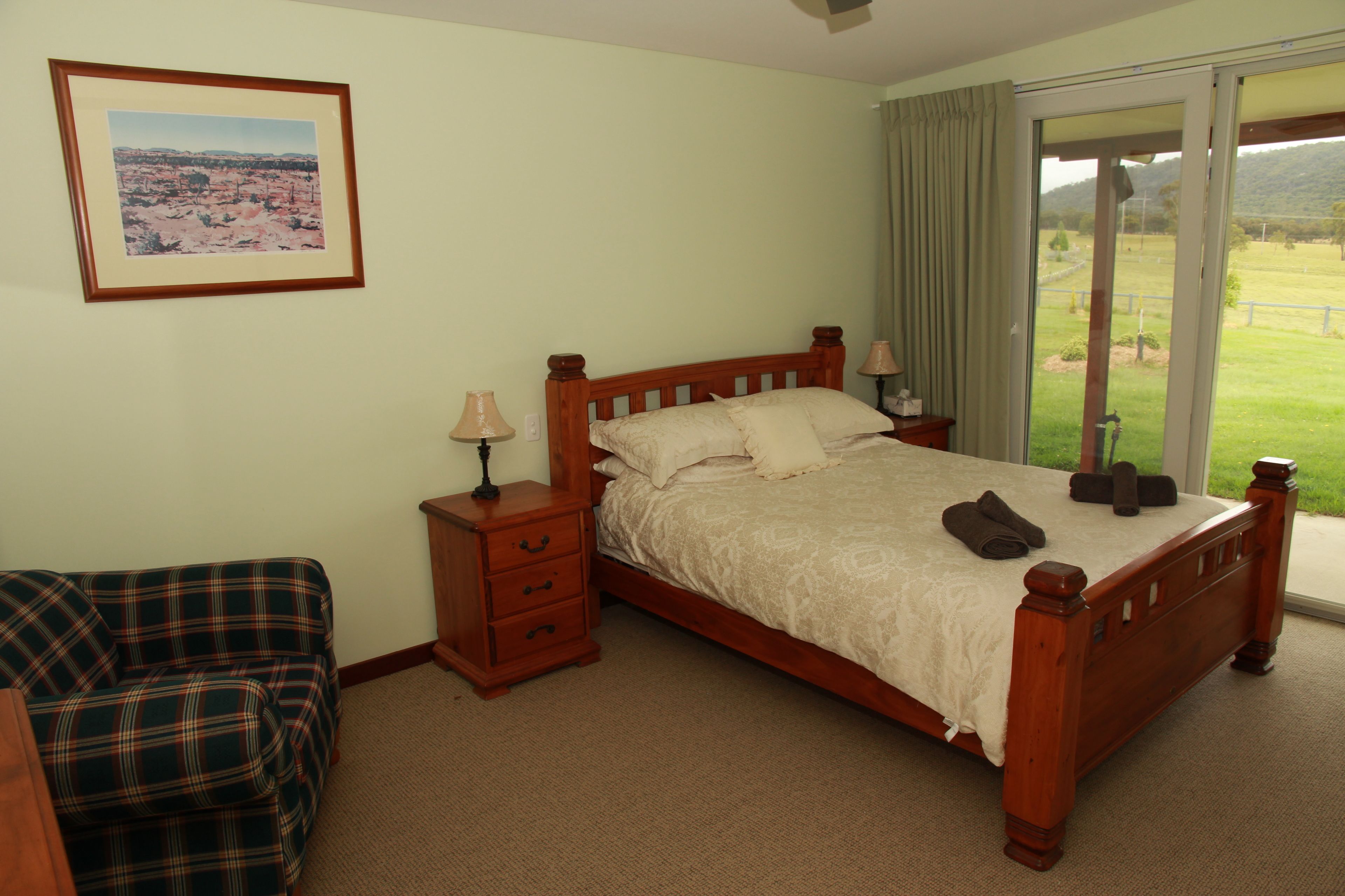 Une chambre à coucher avec un lit en bois, une table de chevet, une lampe, un fauteuil à carreaux et une grande fenêtre donnant sur un paysage verdoyant.
