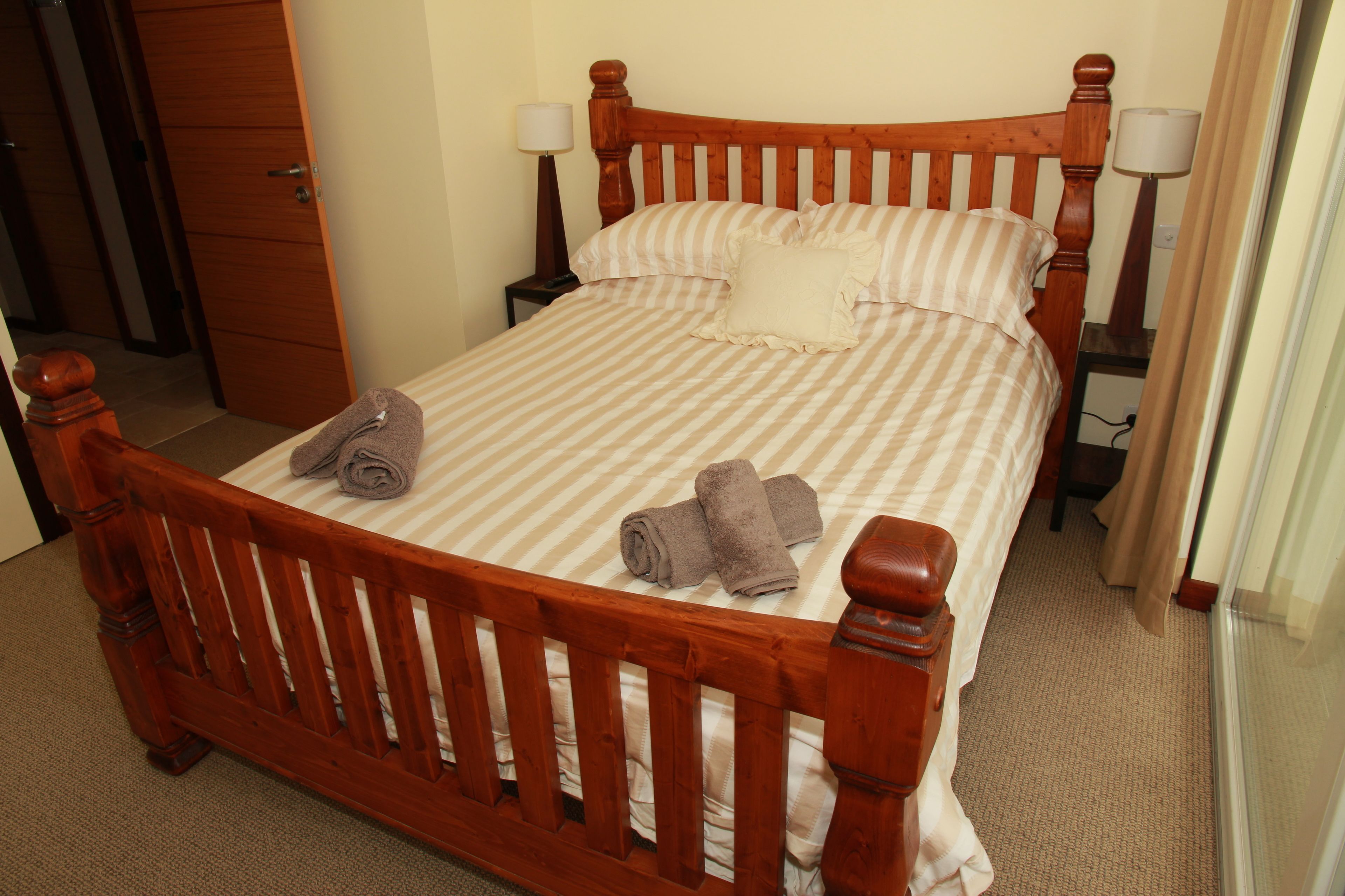 A wooden bed with a striped bedspread and pillows, a wooden nightstand with a lamp, and a rolled towel on the bed.