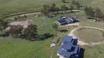 Une vue aérienne d’une propriété rurale comprenant plusieurs bâtiments, dont une grande maison avec un toit bleu, entourée de champs ouverts et de quelques arbres dispersés.