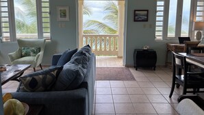 A living room with a sofa, a dining table, and a balcony with a view of palm trees. - On Ocean&Sandy Beach-Private access to beach! Air conditioned. Standby Generator (Rincon)