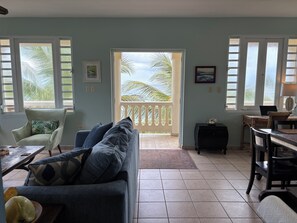 A living room with a sofa, a dining table, and a balcony with a view of palm trees. - On Ocean&Sandy Beach-Private access to beach! Air conditioned. Standby Generator (Rincon)