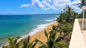 A beach with clear turquoise water, palm trees, and a white building with a balcony. - On Ocean&Sandy Beach-Private access to beach! Air conditioned. Standby Generator (Rincon)
