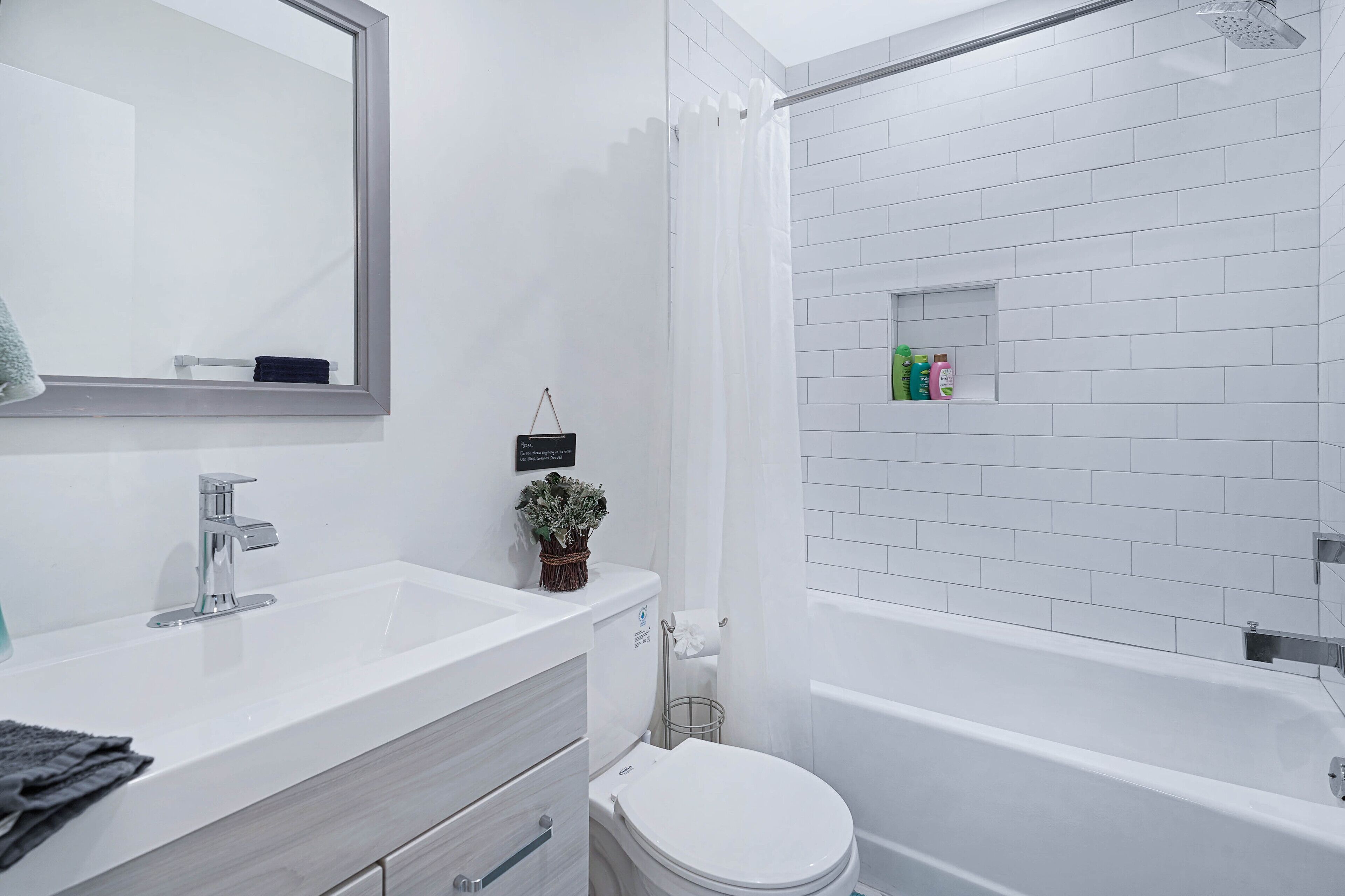 Ein modernes Badezimmer mit einem weißen Waschbecken, einem Spiegel und einer Toilette. Eine Duschkabine mit weißem Vorhang und ein Ablagebereich mit Pflegeprodukten.