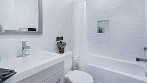 A modern bathroom with a white sink, mirror, and toilet. There is a shower area with a white curtain and a shelf with toiletries. - Stylish Modern Home ★ Heart of Hollywood & Walkable ★ 2 Private Car Spots (Los Angeles)