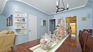 A dining area with a wooden table, striped tablecloth, and a centerpiece of seashells. There is a white door, a white shelf with decorative items, and a ceiling light fixture. - A PERFECT BEACH COTTAGE  Exclusive Private Retreat - STEPS TO BEACH AND BAY (San Diego)