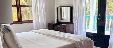 Een slaapkamer met een bed, nachtkastje, lamp, airconditioning en een plafondventilator, met uitzicht op het groen buiten.