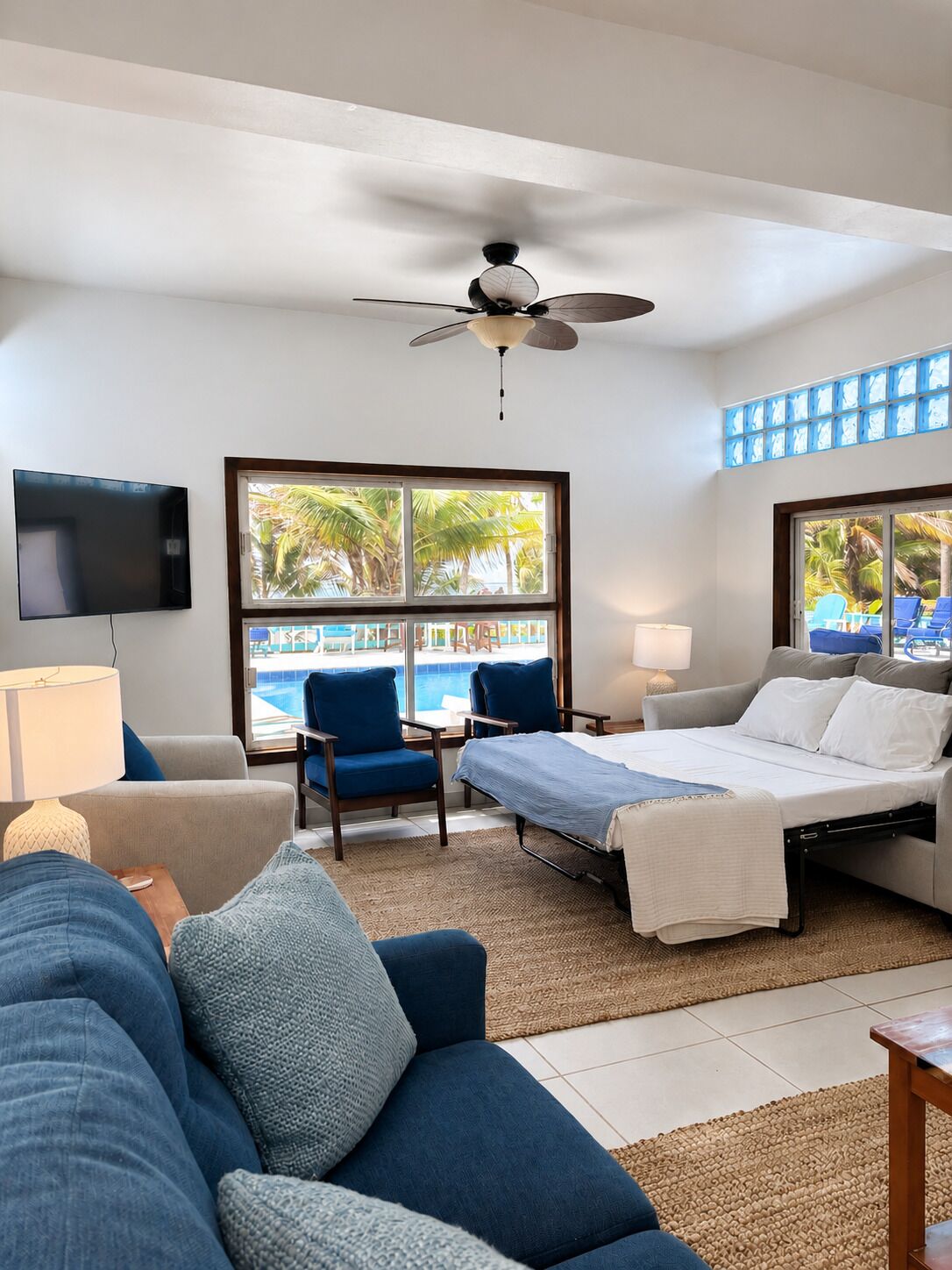 A hotel room with a bed, a sofa, a TV, a ceiling fan, and a window with a view of a pool.