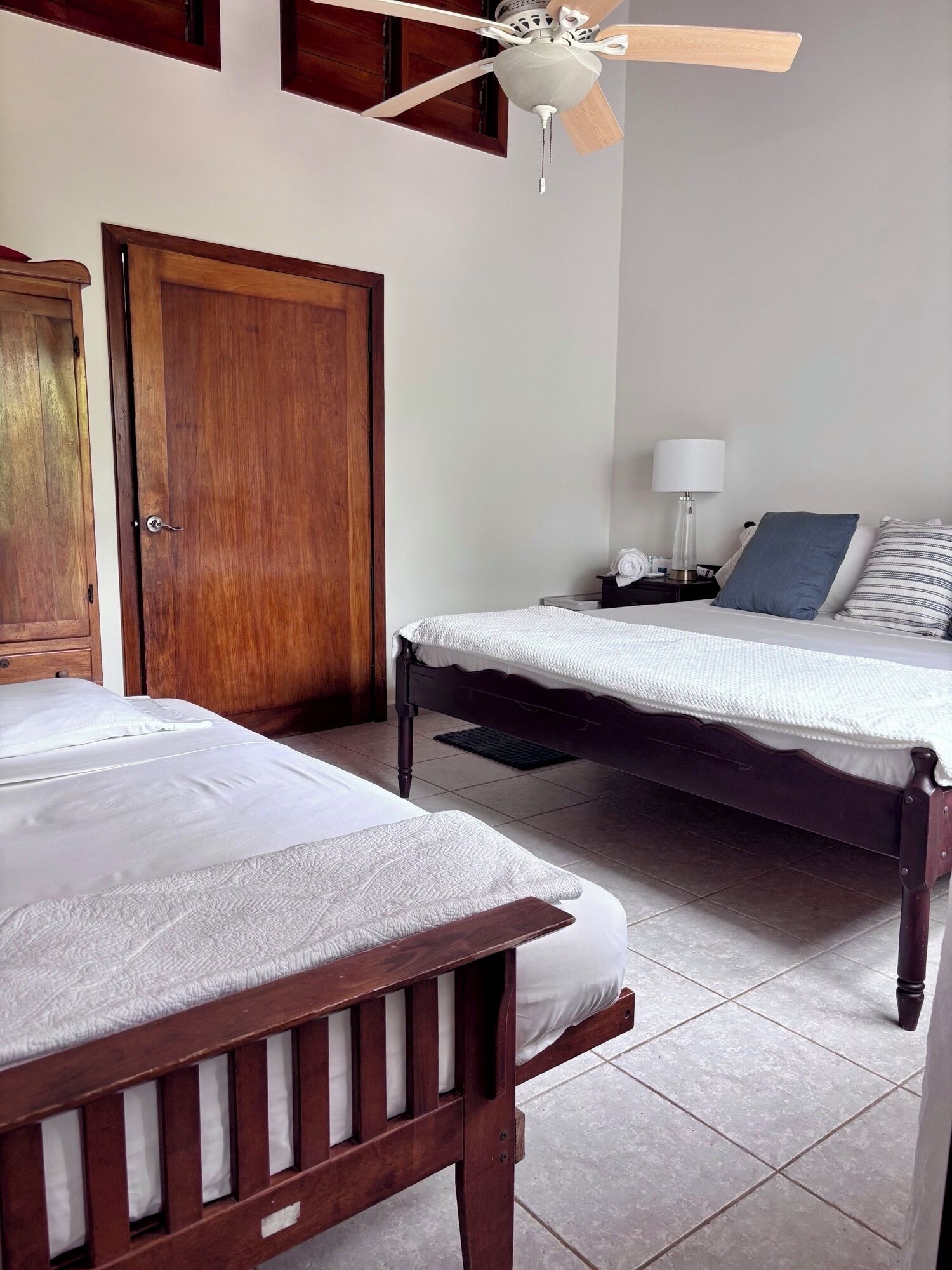 Habitación de hotel con dos camas de madera, un ventilador de techo, una mesita de noche con lámpara y un armario de madera.