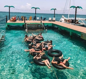 A pool area with people floating on tubes, a wooden deck, and a boat docked nearby.