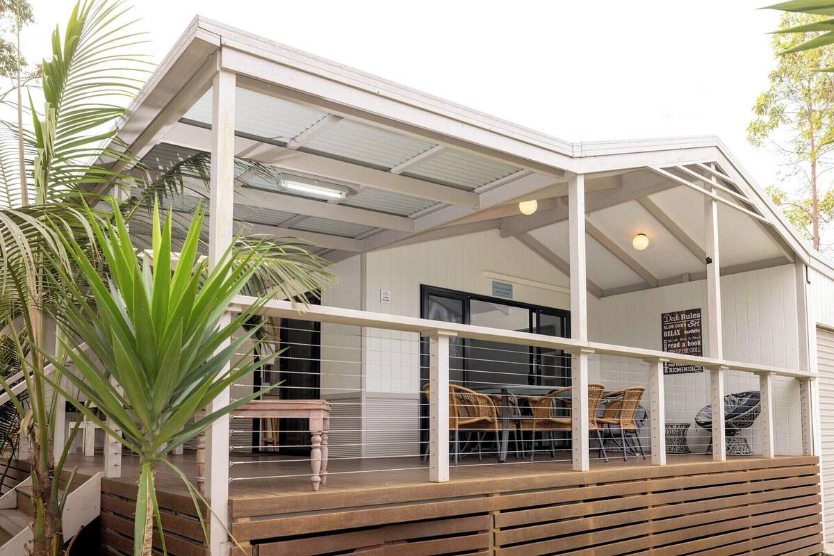 Een modern terras buiten met glazen hekwerk en houten vloer, omgeven door weelderige beplanting.