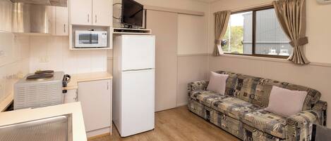 A compact kitchen with a microwave, refrigerator, and a small TV on a shelf. There's a patterned sofa and a window with curtains.