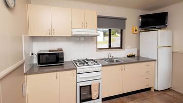 Cocina funcional con microondas, estufa, horno y refrigerador. Se observa una ventana con vista al exterior, un reloj en la pared y un televisor de pantalla plana sobre el refrigerador.